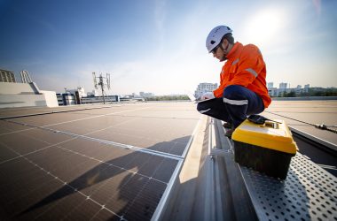 Technician inspecting solar panels on a rooftop in a modern urban environment A technician in an orange jacket and helmet crouches on a rooftop, examining solar panels while holding a clipboard. The city skyline is visible under a clear sky.